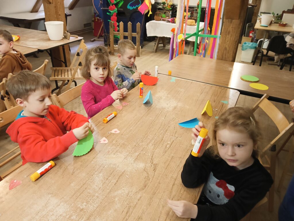 Zdjęcie przedstawia dzieci siedzące przy stole, na którym znajduje się kolorowy papier i kleje. Dzieci robią prace plastyczne.