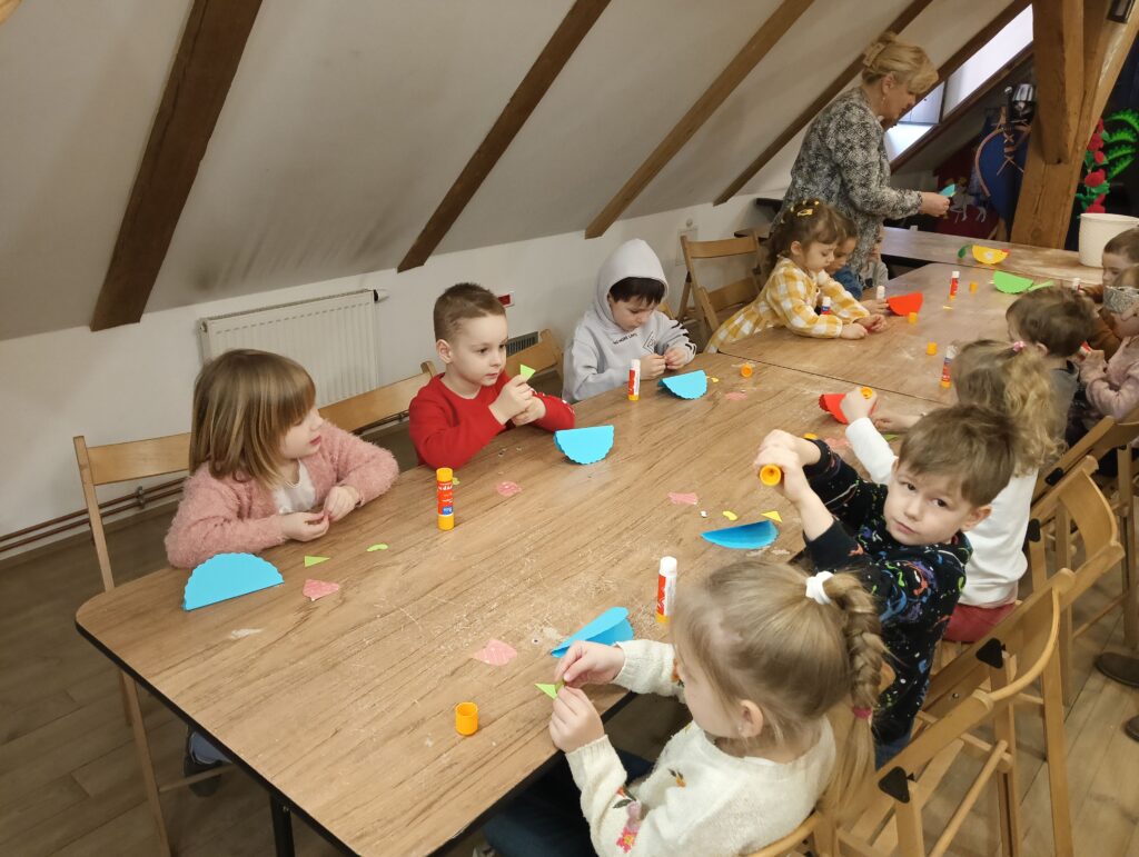 Zdjęcie przedstawia dzieci siedzące przy stole. Pani nauczycielka rozdaje dzieciom kolorowy papier i kleje, które posłużą do zrobienia prac plastycznych.