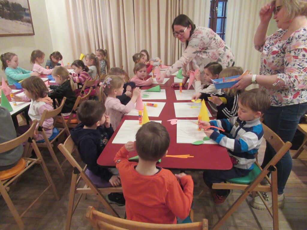 Zdjęcie grupy dzieci z opiekunami. Dzieci siedzą przy stołach, wykonują prace plastyczne.