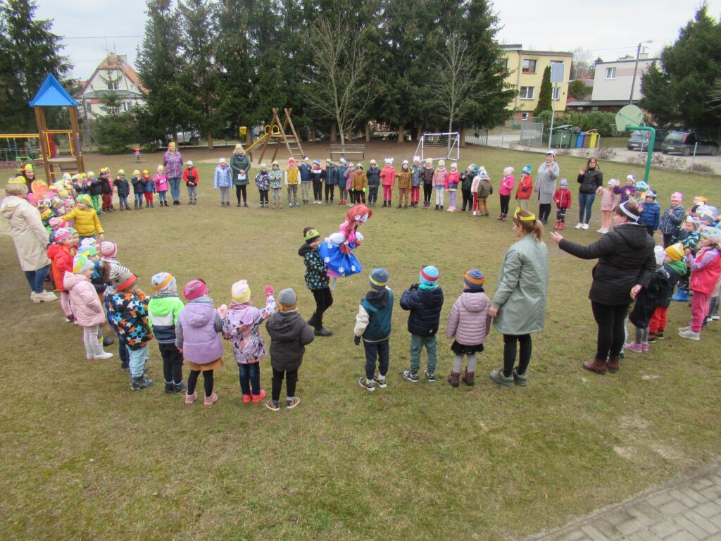 Zdjęcie przedstawia przedszkolaków stojących na placu przedszkolnym w kole wraz z nauczycielami.