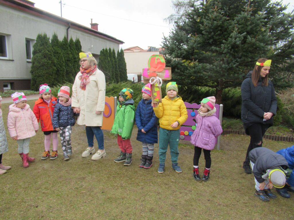 Zdjęcie przedstawia grupę dzieci stojących na placu przedszkolnym wraz z nauczycielem i opiekunem. Jedno z dzieci trzyma w rękach Marzannę.