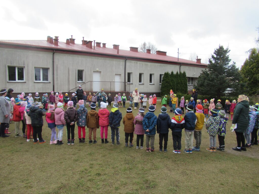 Zdjęcie przedstawia przedszkolaków stojących na placu przedszkolnym w kole wraz z nauczycielami.