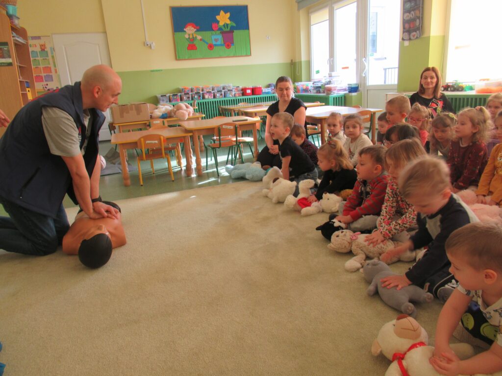 Zdjęcie przedstawia dzieci i opiekunów siedzących na dywanie w sali przedszkolnej. Dzieci mają ze sobą maskotki. Obok jest pan, który pokazuje jak robić uciski na fantomie.