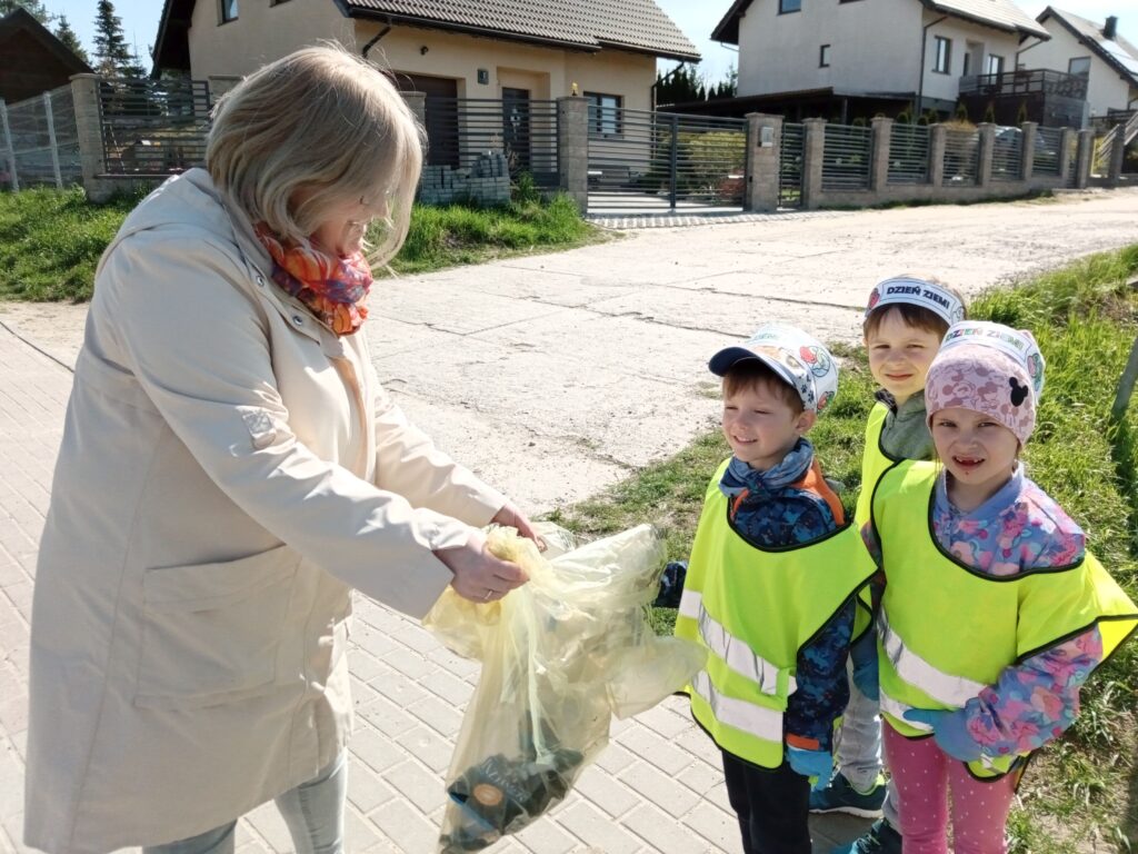 Zdjęcie przedstawia troje dzieci w kamizelkach odblaskowych w plenerze wraz z panią nauczycielką.