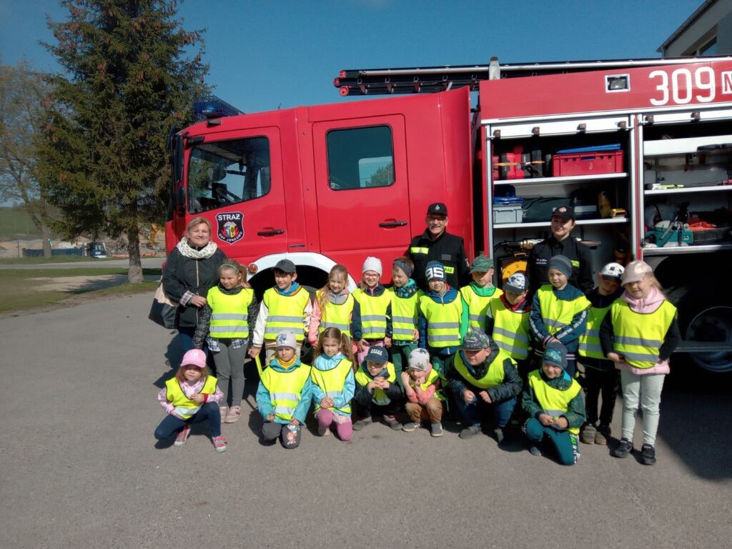 Zdjęcie przestawia grupę dzieci wraz z opiekunem i strażakami na tle wozu strażackiego.