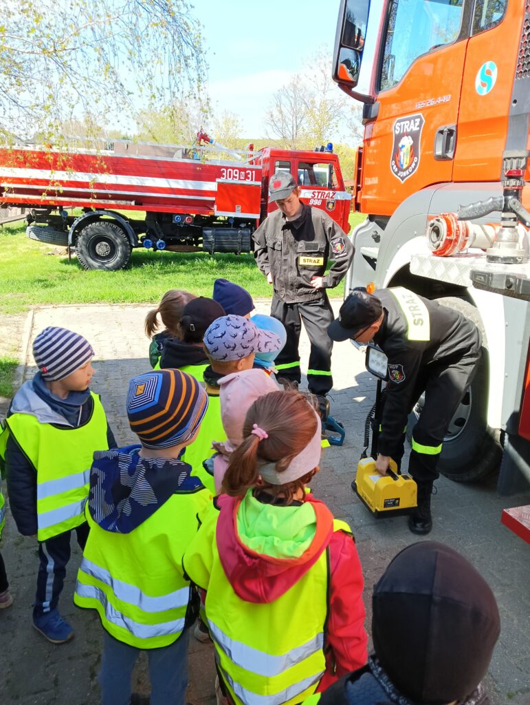 Zdjęcie przestawia grupę dzieci i strażaków. Obok stoją wozy strażackie.