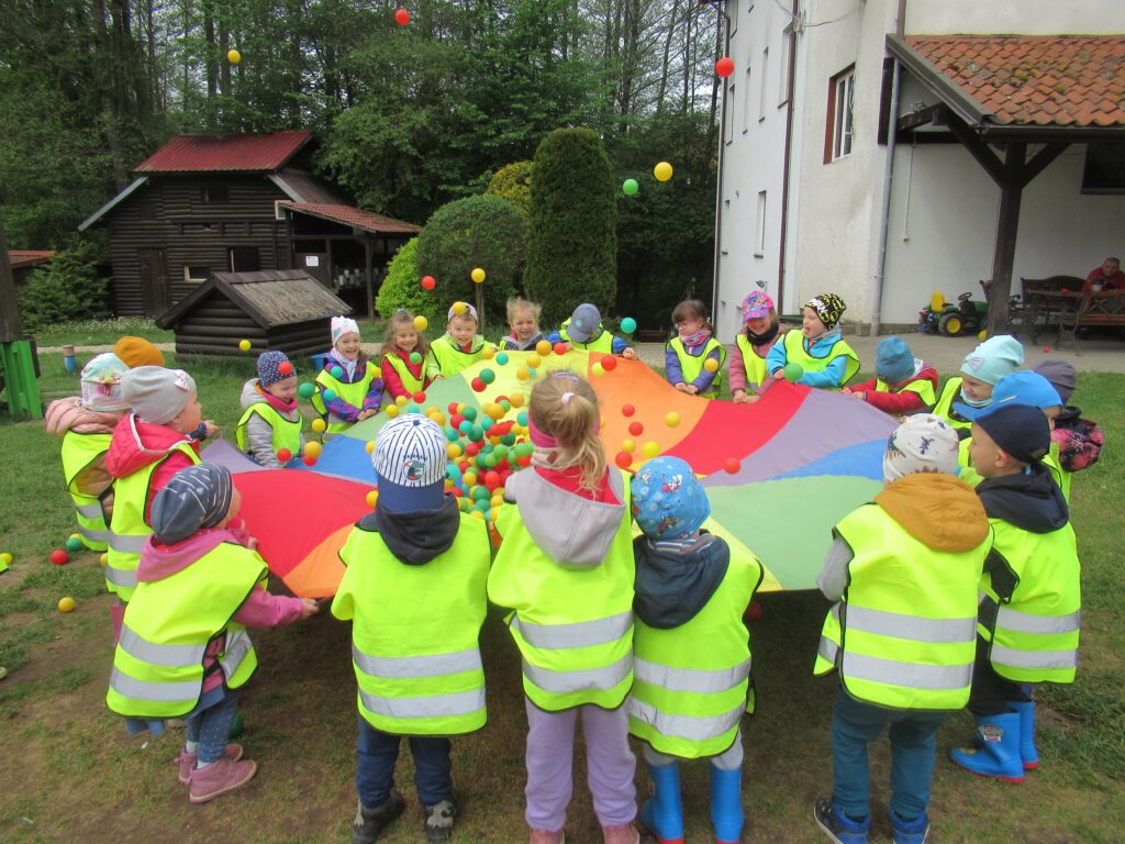 Zdjęcie przedstawia grupę dzieci na placu zabaw. Dzieci stoją w kole, bawią się piłeczkami na chuście animacyjnej.