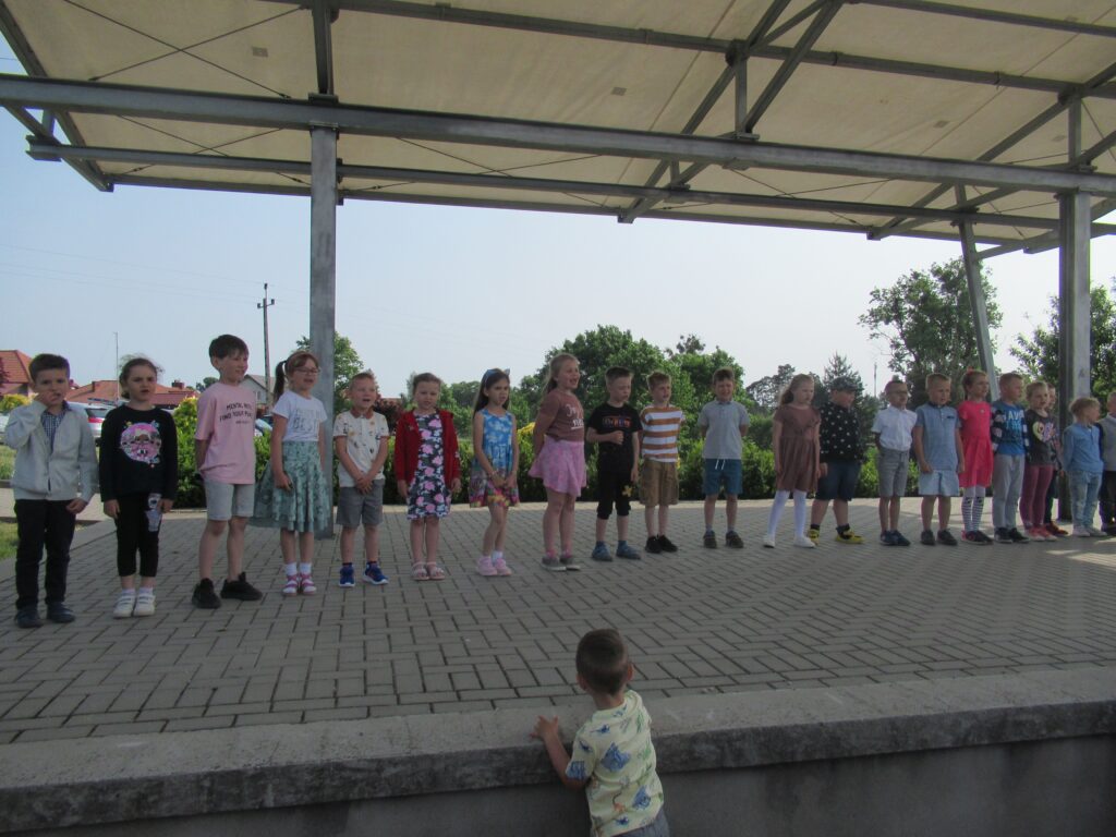 Zdjęcie przedstawia grupę dzieci, którzy występują na scenie. Z tyłu zdjęcia widać krzewy.