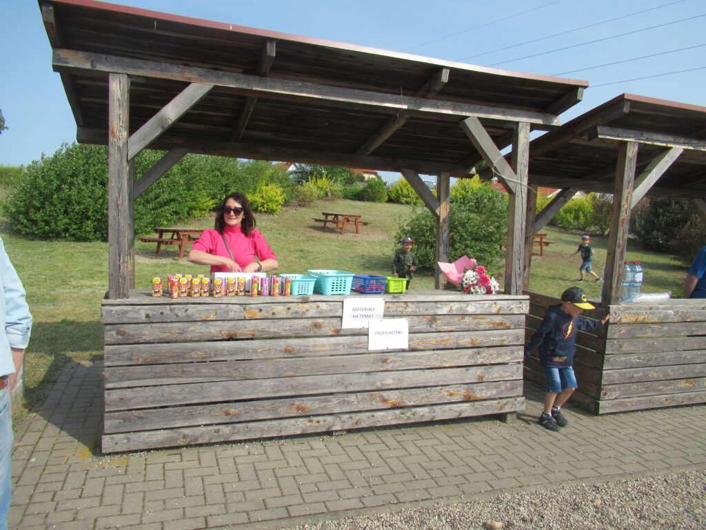Zdjęcie przedstawia jedna panią stojącą pod wiatą z wystawką soków i materiałów w koszykach plastikowych oraz dzieci. Z tyłu wiaty widać drzewa oraz stoliki z ławkami.