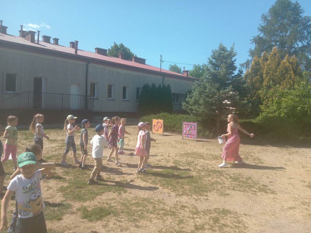 Zdjęcie przedstawia grupę dzieci wraz z panią bawiących się na placu zabaw. Z tyłu widać budynek przedszkola oraz drzewa.