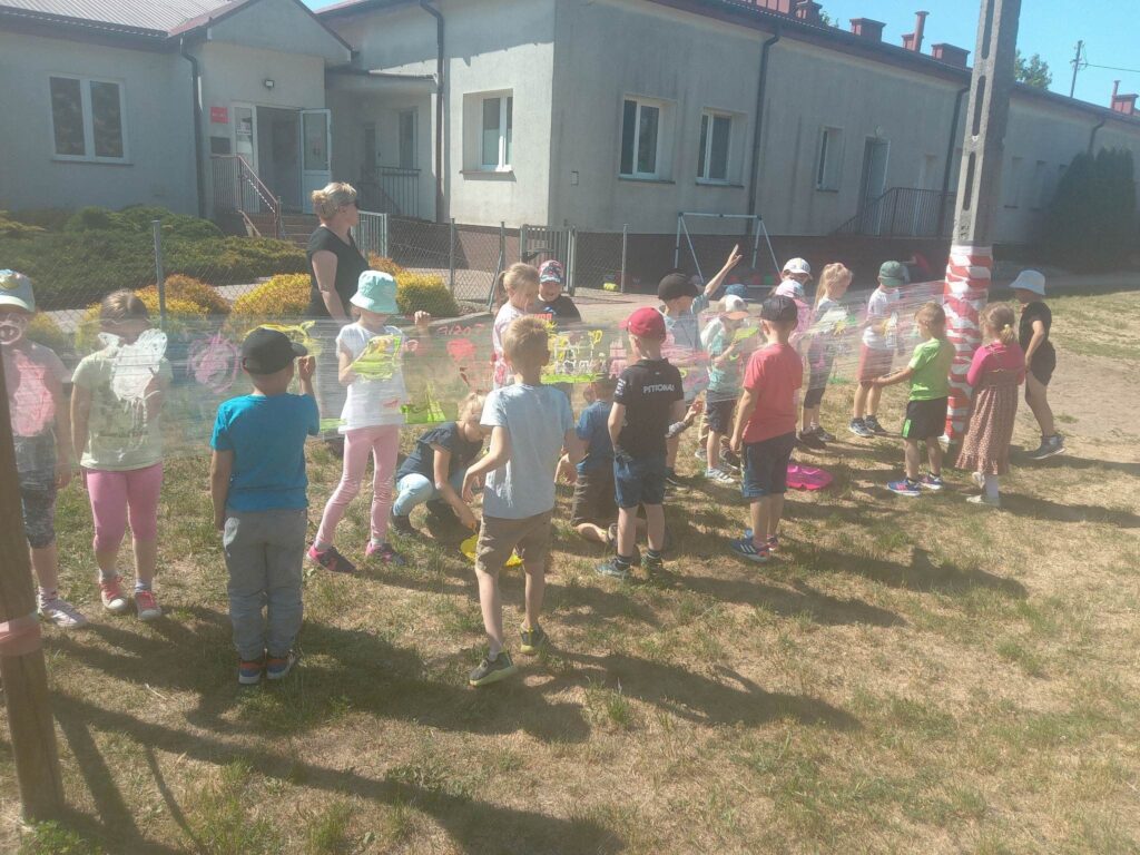 Zdjęcie przedstawia grupę dzieci wraz z panią bawiących się na placu zabaw. Z tyłu widać budynek przedszkola.