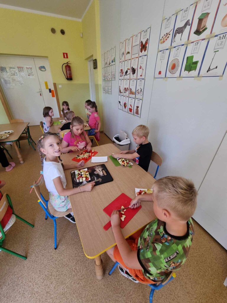 Zdjęcie przedstawia grupę dzieci siedzących przy stolikach w sali przedszkolnej.
