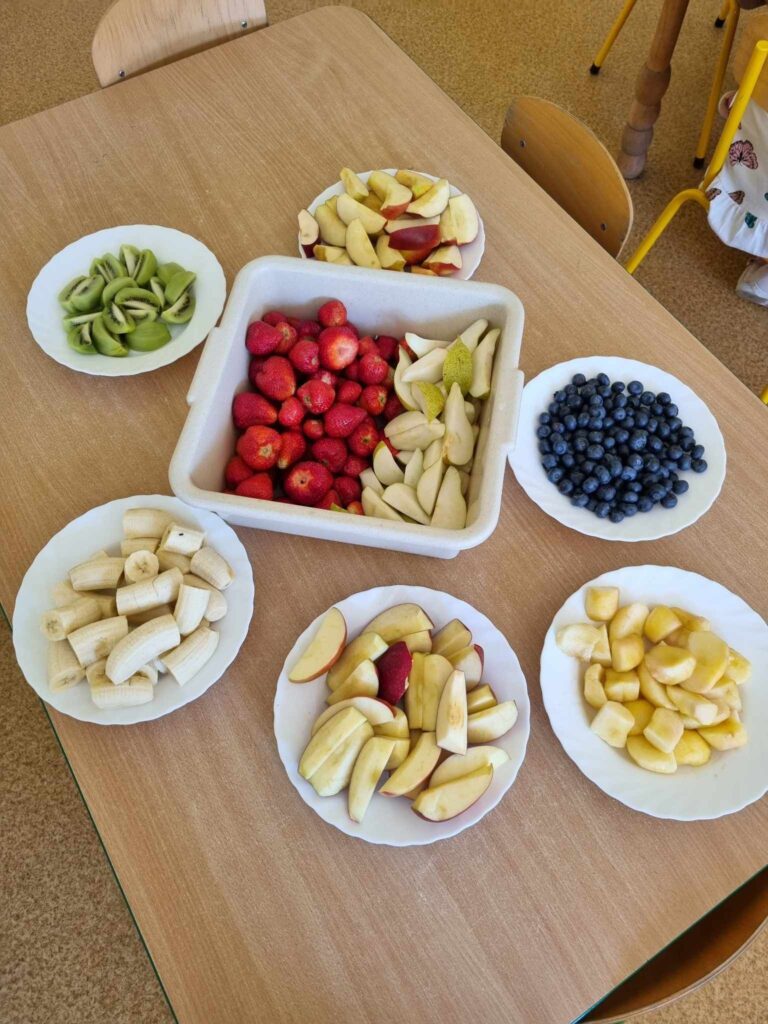 Zdjęcie przedstawia pokrojone owoce na kilku talerzach w sali przedszkolnej.