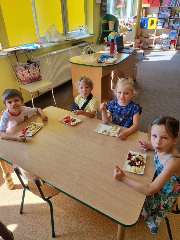 Zdjęcie przedstawia grupę dzieci siedzących przy stoliku w sali przedszkolnej.