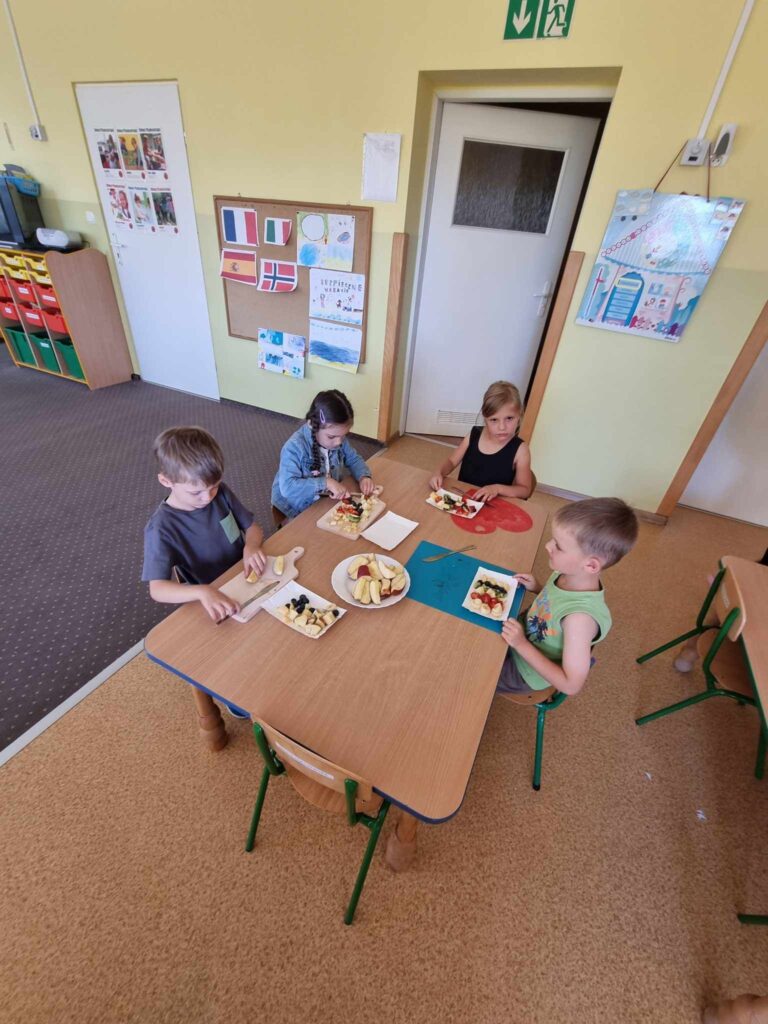Zdjęcie przedstawia grupę dzieci siedzących przy stoliku w sali przedszkolnej.