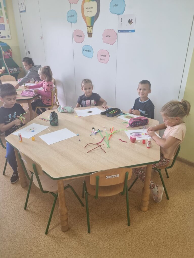 Zdjęcie przedstawia grupę dzieci w sali przedszkolnej, siedzą przy stolikach robią prace plastyczne.