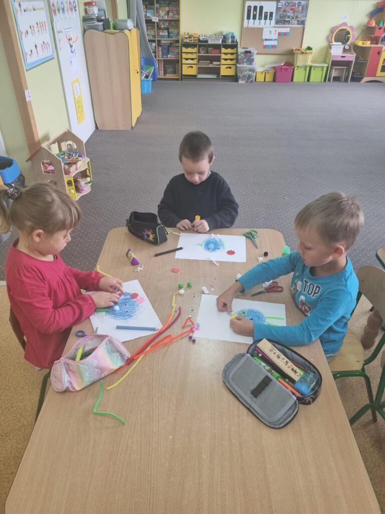 Zdjęcie przedstawia dzieci w sali przedszkolnej, siedzą przy stoliku robią prace plastyczne.