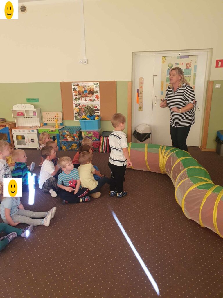 Zdjęcie przedstawia panią i dzieci bawiące się w sali przedszkolnej.