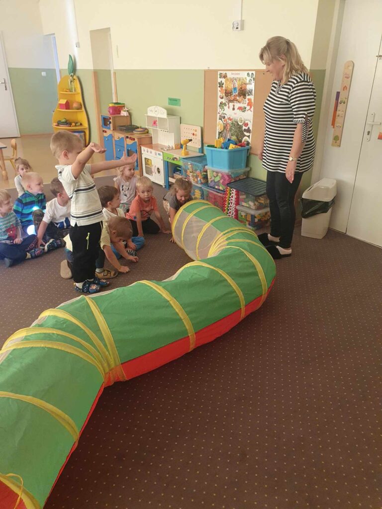 Zdjęcie przedstawia panią i grupę dzieci bawiących się w sali przedszkolnej.