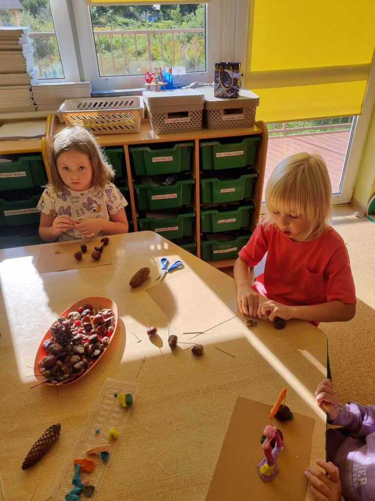Zdjęcie przedstawia dzieci siedzące przy stoliku w sali przedszkolnej - tworzą prace plastyczne.