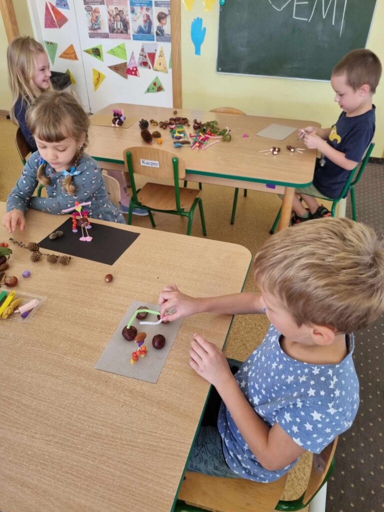 Zdjęcie przedstawia dzieci siedzące przy stolikach w sali przedszkolnej - tworzą prace plastyczne.