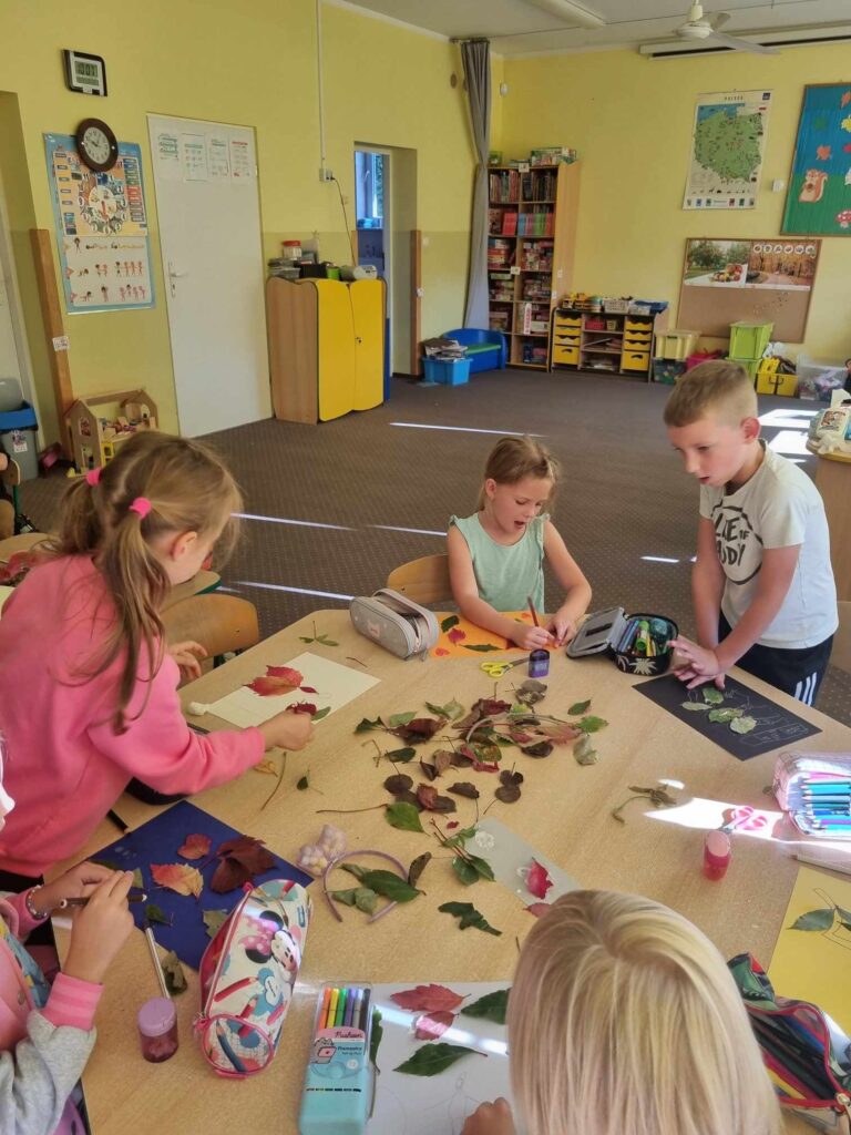 Zdjęcie przedstawia dzieci zgromadzone przy stoliku w sali przedszkolnej - tworzą prace plastyczne.