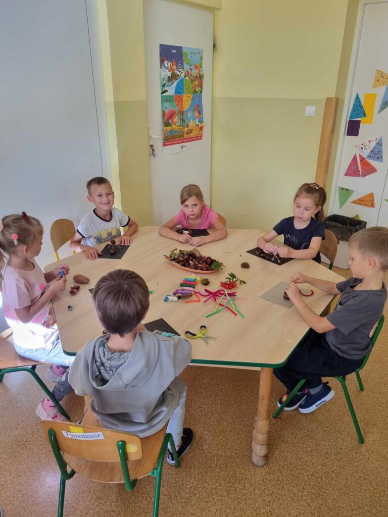 Zdjęcie przedstawia dzieci siedzące przy stoliku w sali przedszkolnej - tworzą prace plastyczne.