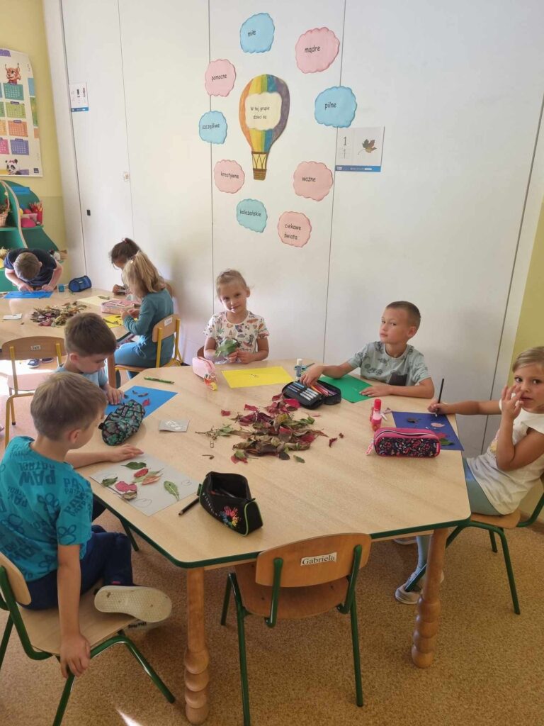 Zdjęcie przedstawia dzieci siedzące przy stolikach w sali przedszkolnej - tworzą prace plastyczne.