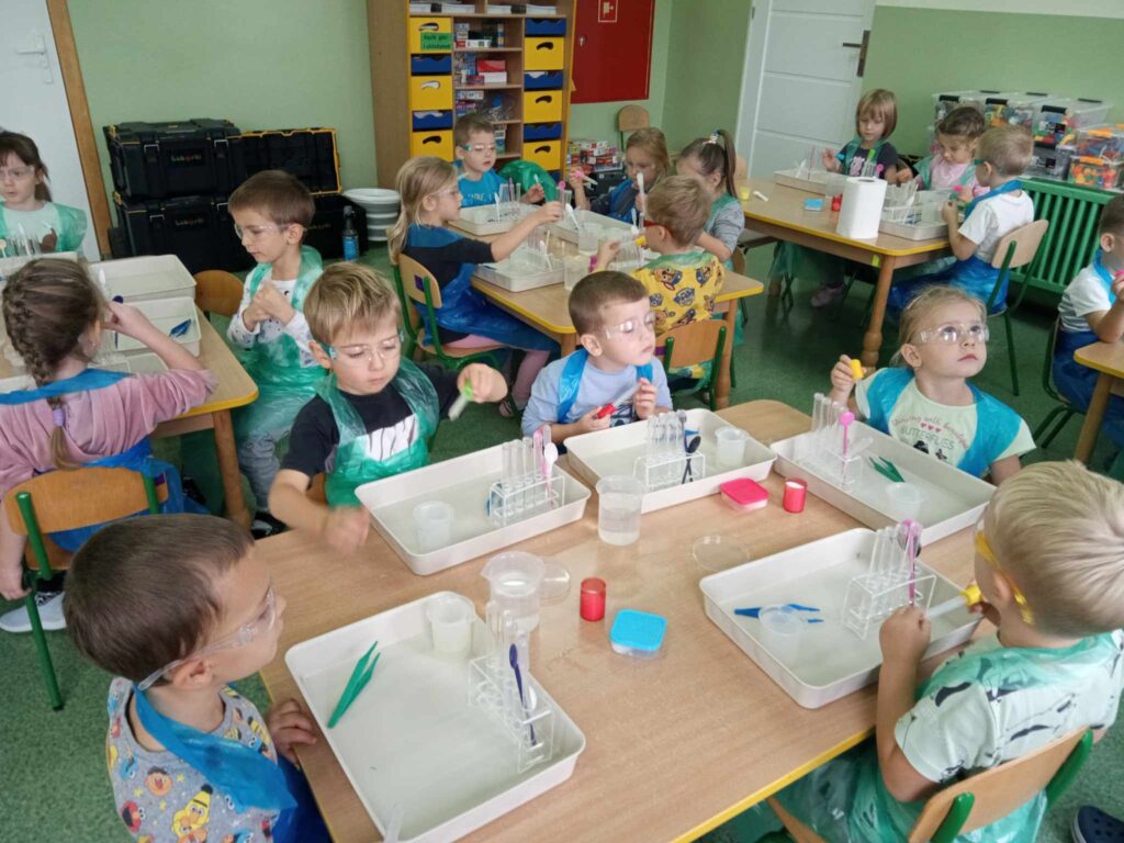 Zdjęcie przedstawia dzieci siedzące przy stolikach w sali przedszkolnej.