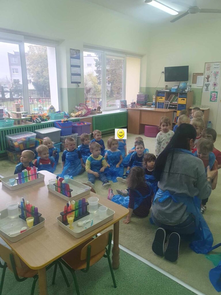 Zdjęcie przedstawia panią oraz grupę dzieci w sali przedszkolnej.