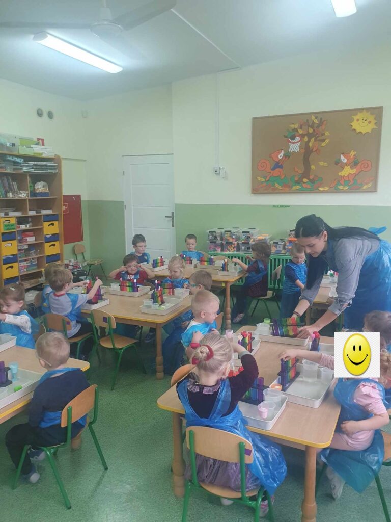 Zdjęcie przedstawia panią oraz dzieci siedzące przy stolikach w sali przedszkolnej.