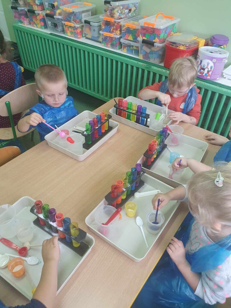 Zdjęcie przedstawia dzieci siedzące przy stoliku w sali przedszkolnej.