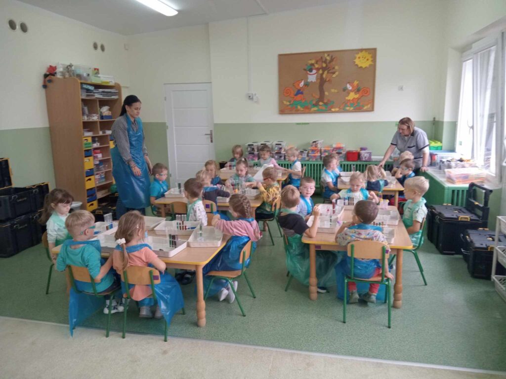 Zdjęcie przedstawia panie oraz dzieci siedzące przy stolikach w sali przedszkolnej.
