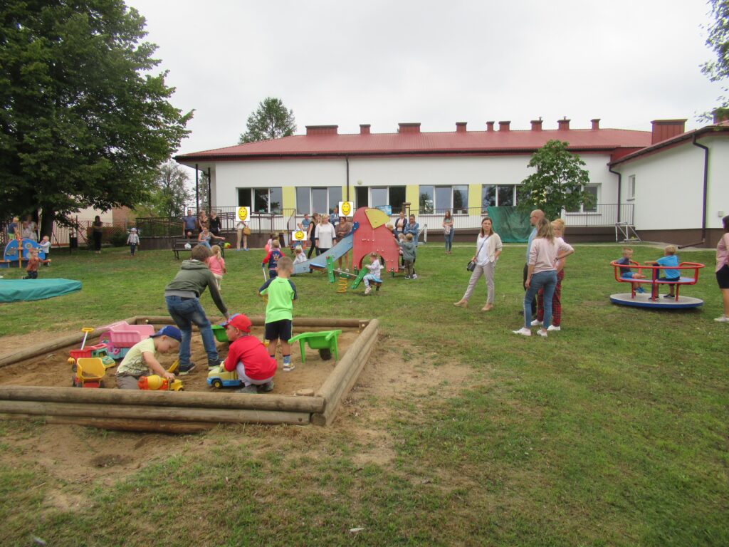 Zdjęcie przedstawia grupę ludzi w ogrodzie przedszkolnym. Z tyłu widać budynek przedszkola.