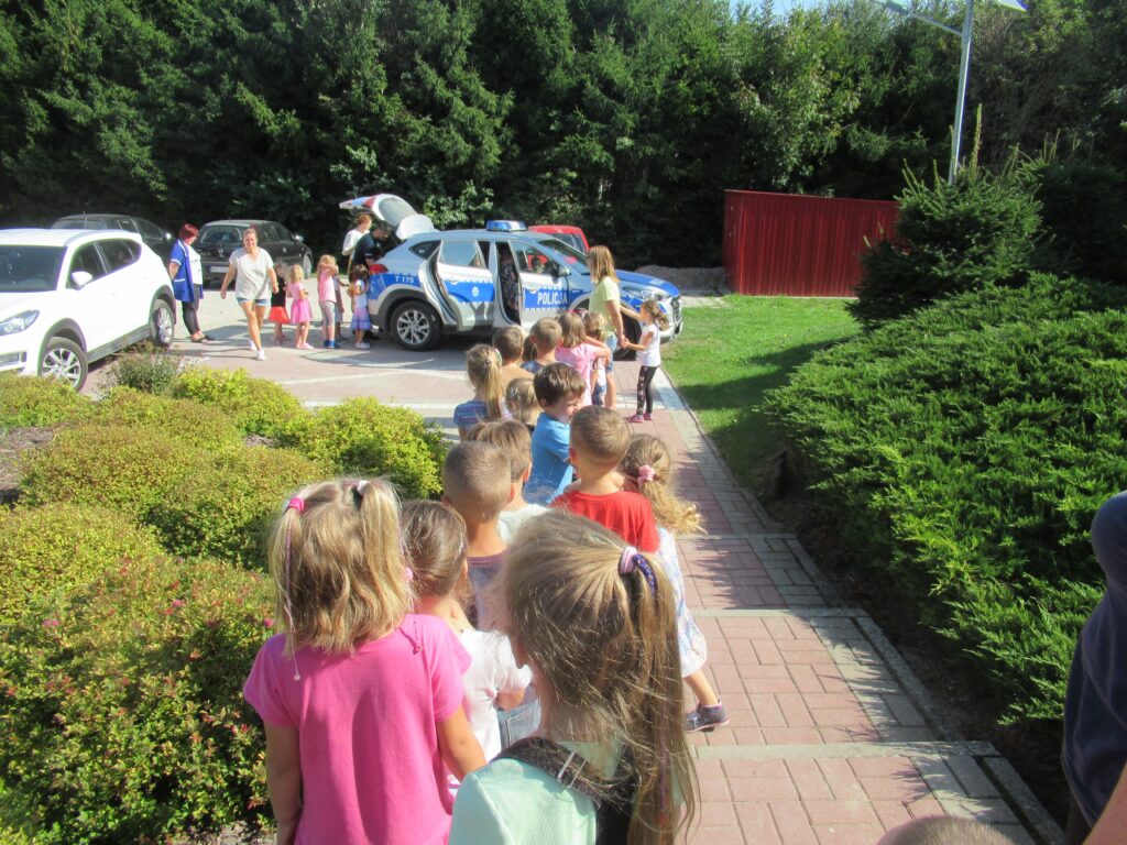 Zdjęcie przedstawia grupę ludzi na parkingu przedszkolnym wraz z samochodami.