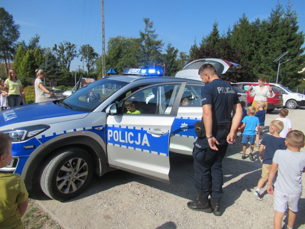 Zdjęcie przedstawia grupę ludzi na parkingu przedszkolnym wraz z samochodami.