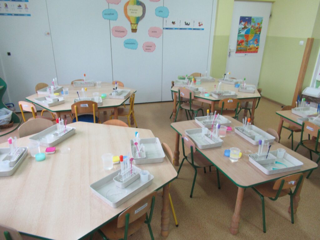 Zdjęcie przedstawia stoliki w sali przedszkolnej. Na stolikach stoją profesjonalne zestawy na tacach do przeprowadzenia badań.