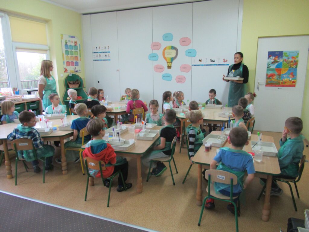 Zdjęcie przedstawia panie oraz dzieci siedzące przy stolikach w sali przedszkolnej.