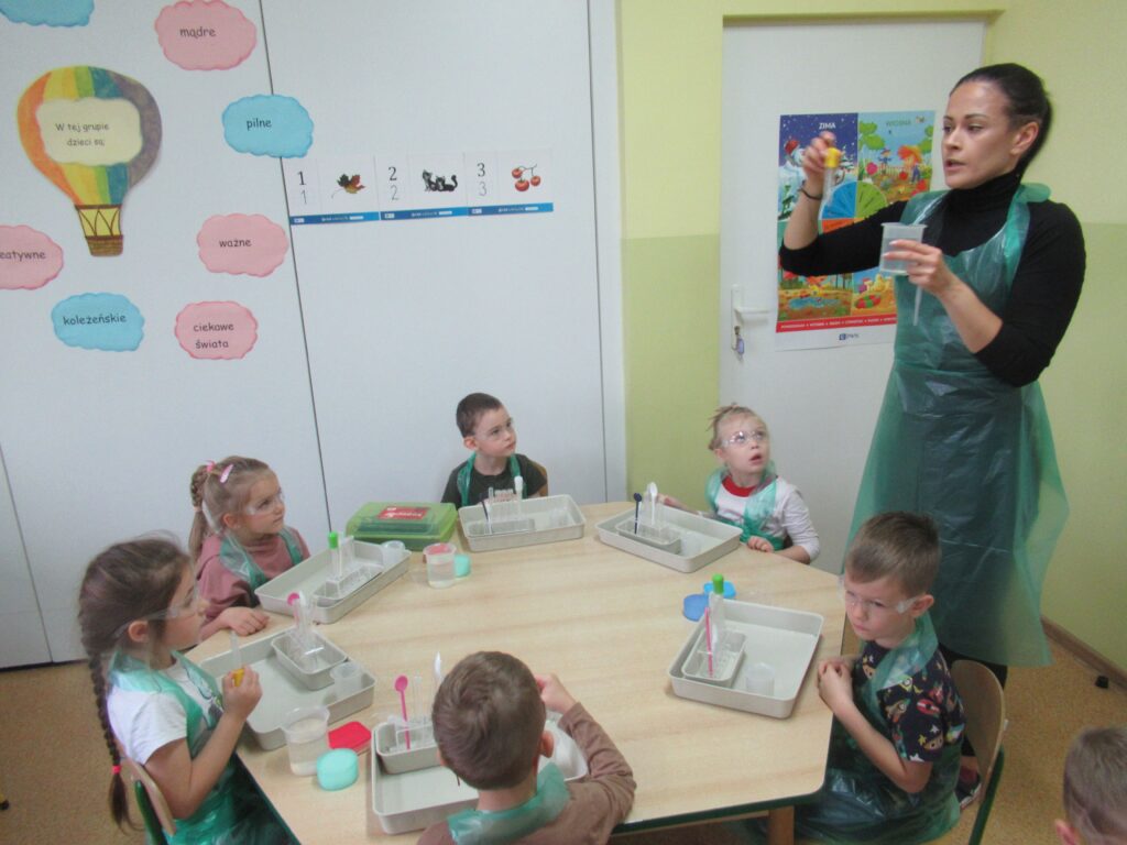 Zdjęcie przedstawia panią oraz dzieci siedzące przy stoliku w sali przedszkolnej.