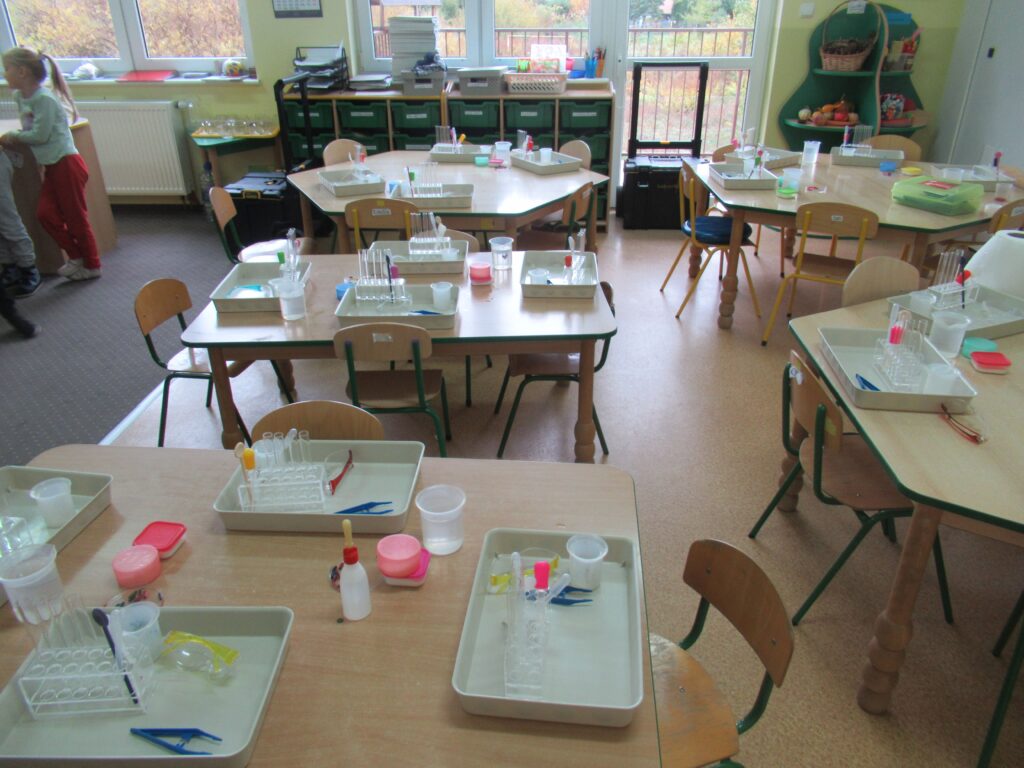 Zdjęcie przedstawia stoliki w sali przedszkolnej. Na stolikach stoją profesjonalne zestawy na tacach do przeprowadzenia badań.