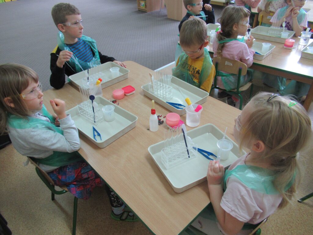 Zdjęcie przedstawia dzieci siedzące przy stolikach w sali przedszkolnej.