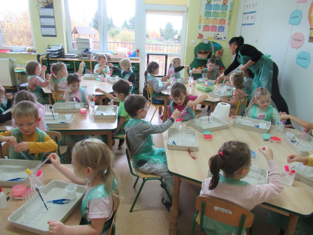 Zdjęcie przedstawia panią oraz dzieci siedzące przy stolikach w sali przedszkolnej.