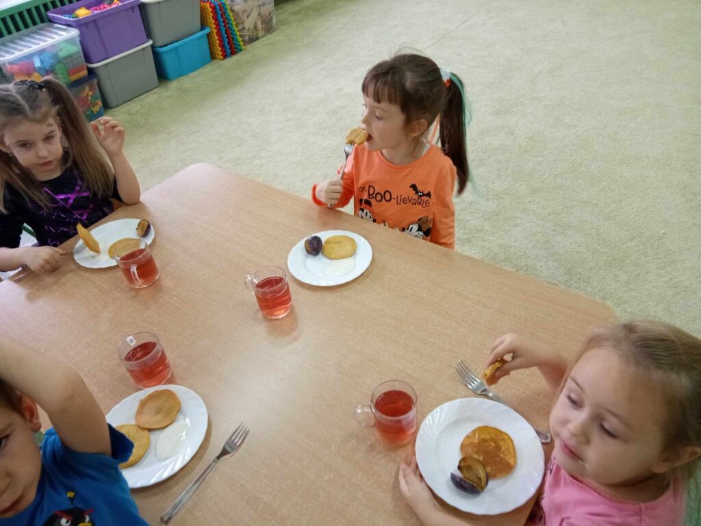 Zdjęcie przedstawia dzieci siedzące przy stolikach, jedzą pyszne racuszki z dyni.