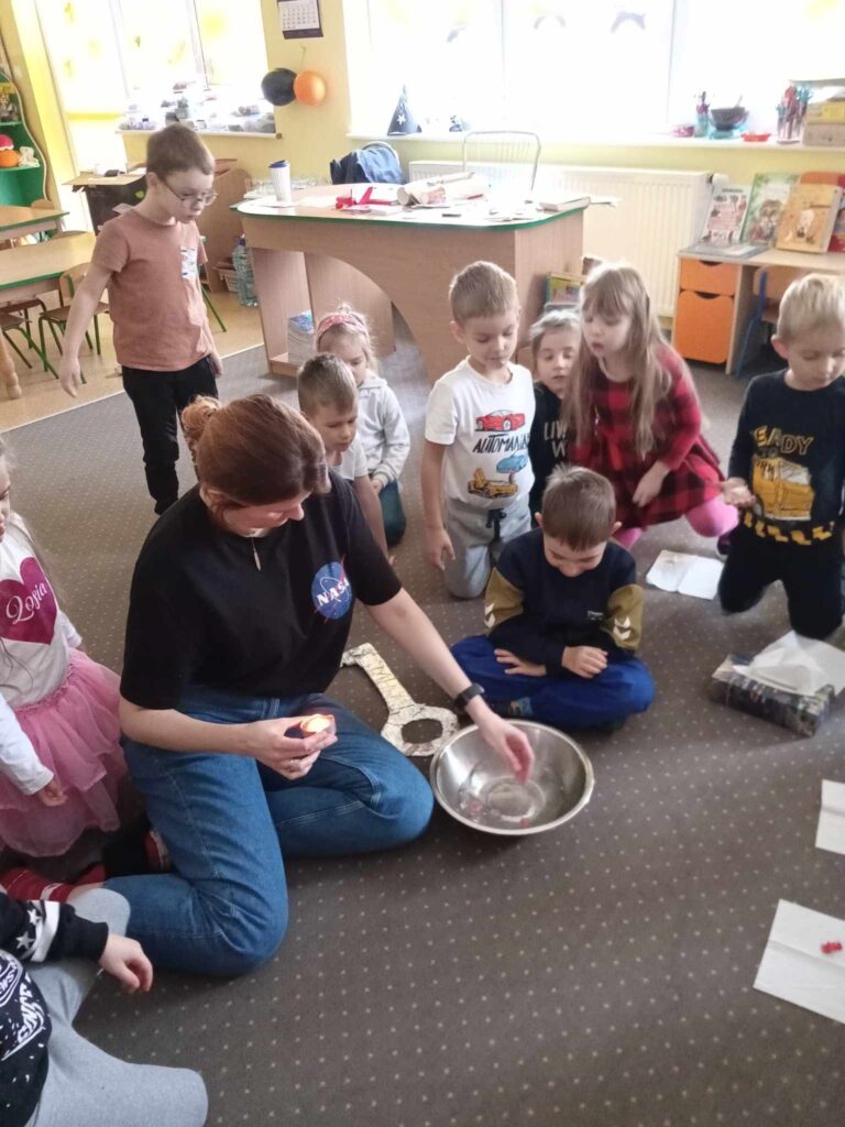 Zdjęcie przedstawia panią oraz grupę dzieci w sali przedszkolnej.