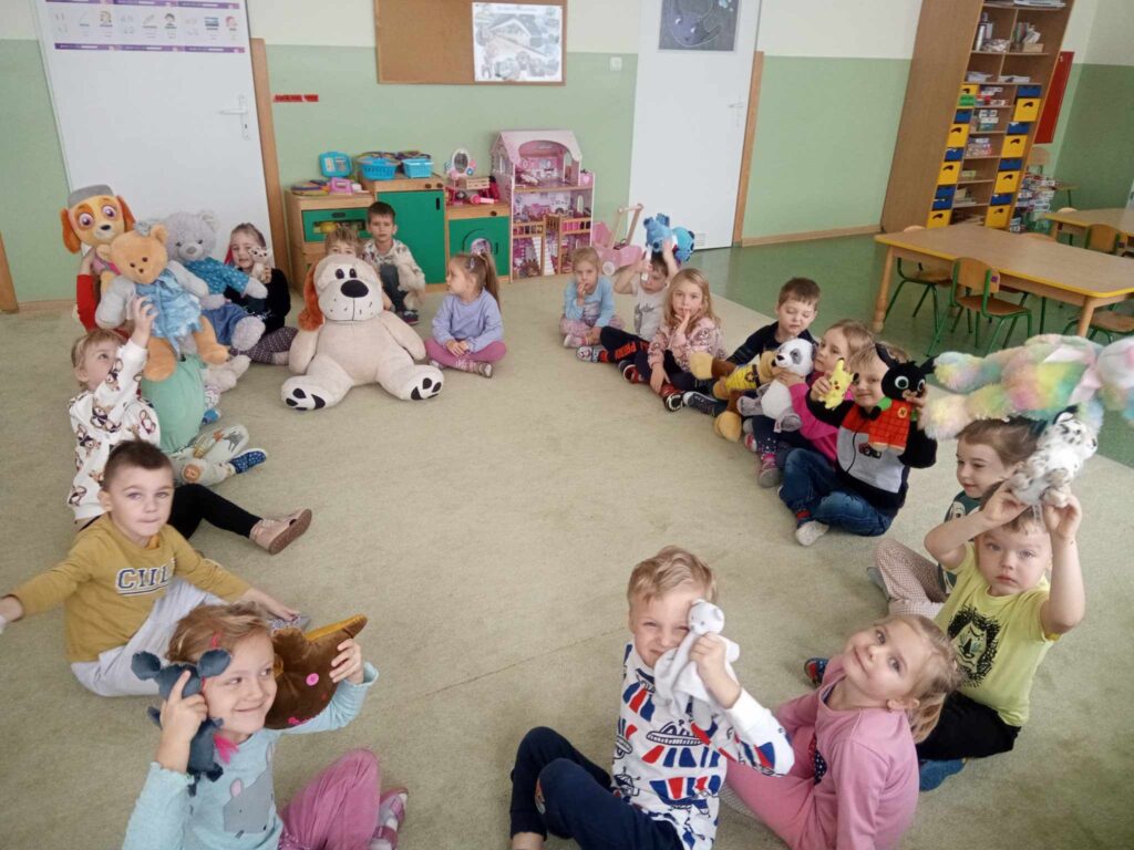 Zdjęcie przedstawia dzieci w sali przedszkolnej siedzące w kole, w rękach trzymają misie.