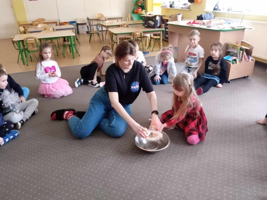 Zdjęcie przedstawia panią oraz grupę dzieci siedzących na wykładzinie w sali przedszkolnej.