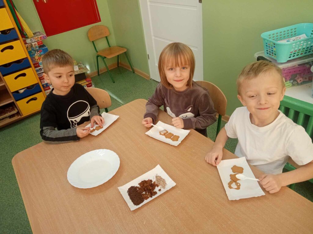 Zdjęcie przedstawia dzieci siedzące przy stolikach w sali przedszkolnej.