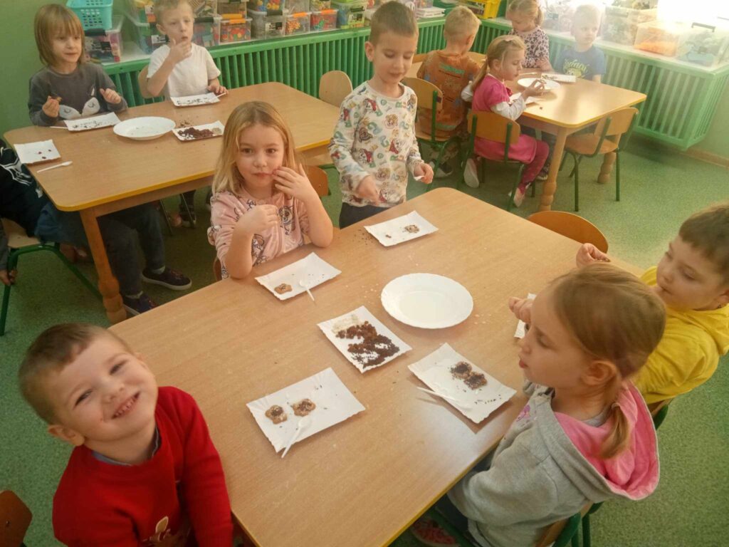 Zdjęcie przedstawia dzieci siedzące przy stolikach w sali przedszkolnej.