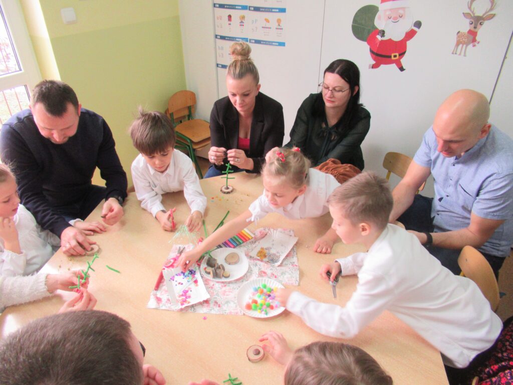 Zdjęcie przedstawia ludzi siedzących przy stoliku w sali przedszkolnej, wykonują prace plastyczne.