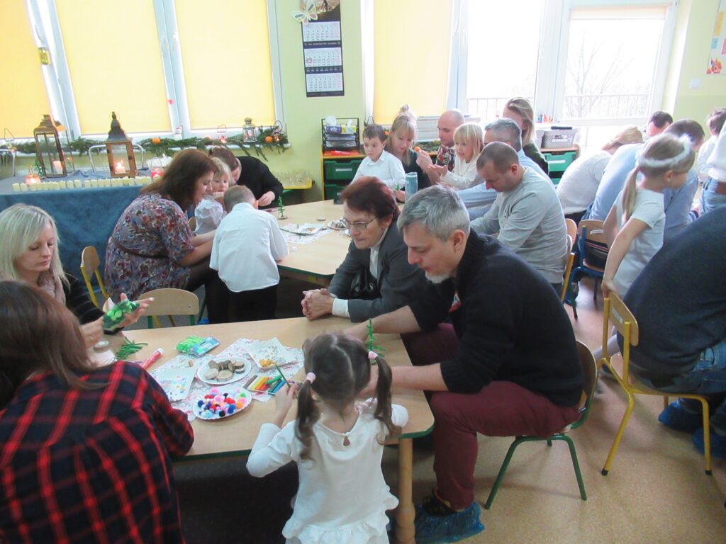 Zdjęcie przedstawia ludzi siedzących przy stolikach w sali przedszkolnej, wykonują prace plastyczne.